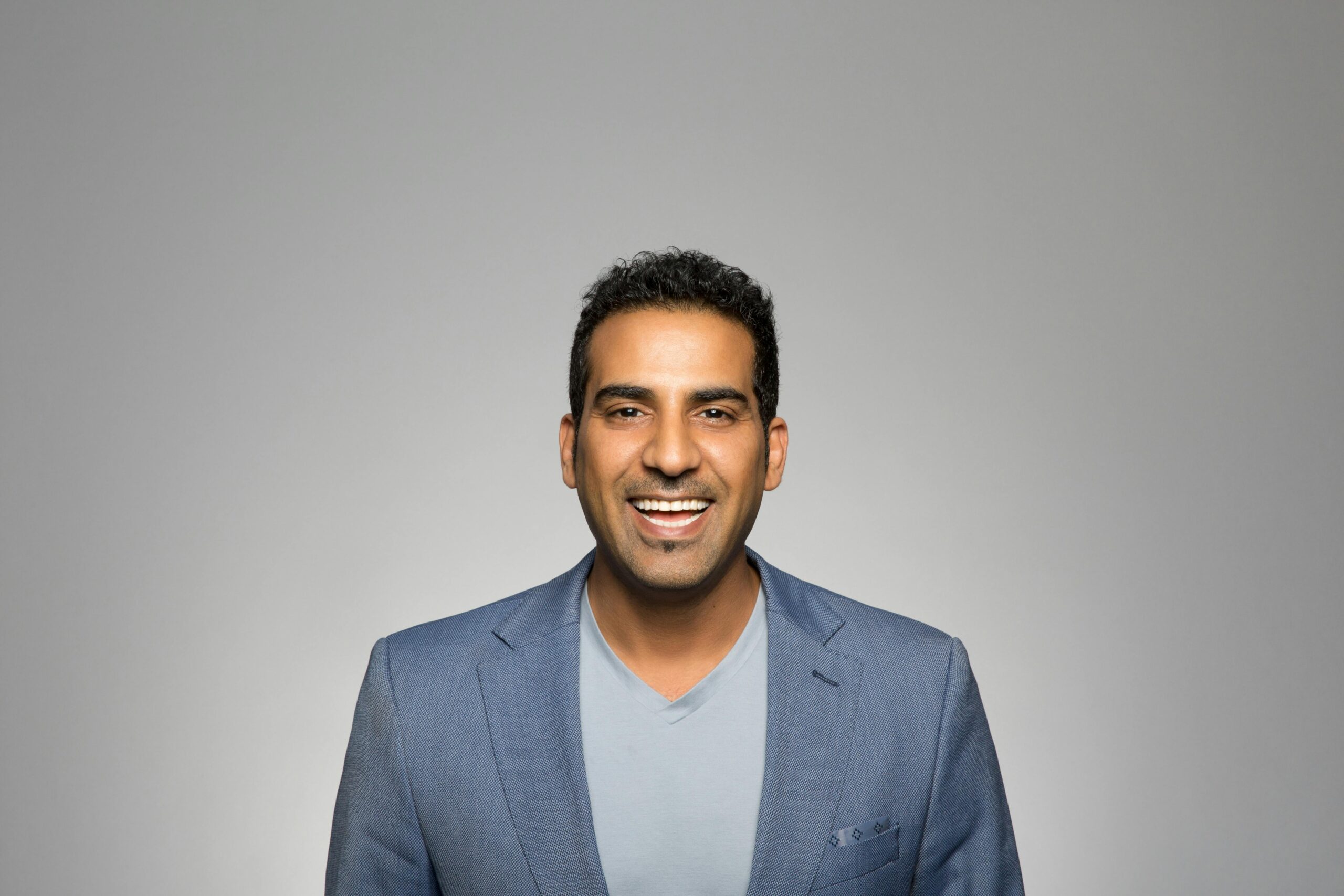 Portrait of a joyful man in a blue blazer, smiling against a plain gray backdrop.